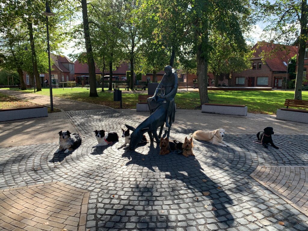 Sieben Hunde liegen rechts und links neben einer Statue mit einem Esel, auf dem ein Mönch sitzt. Die Statue steht auf einem Platz umringt von Sitzgegelenheiten und Grasflächen.